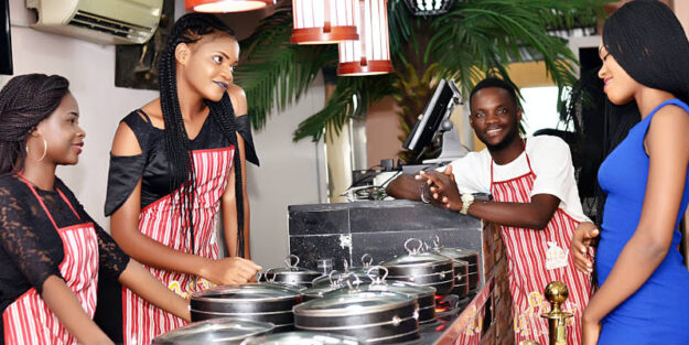 Two Women and Man Wearing Aprons, With a Customer in a Blue Dress in Front of Pots of Food, Corona Estate Planning Attorney.