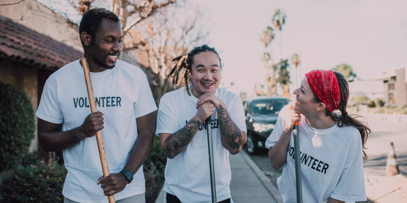 Three individuals, two men and a woman, forming a volunteer tree with shirts that read 'volunteer' and holding broom sticks, symbolizing collaborative efforts in philanthropy.